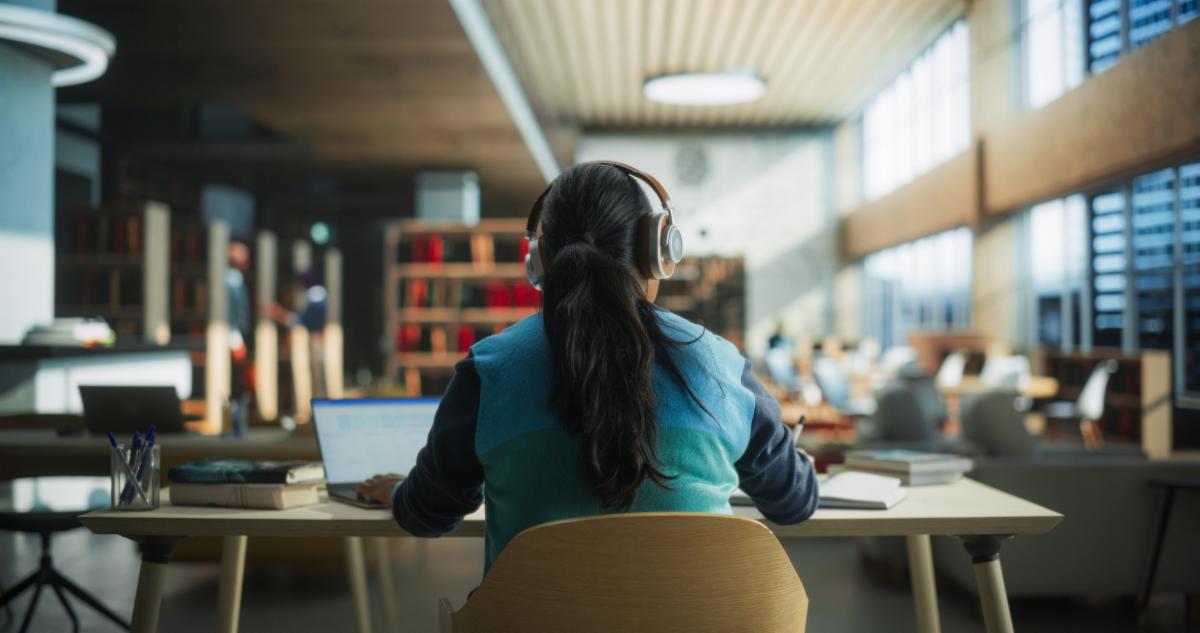 Female Student Wearing Headphones while Working on University Homework in a Public Library. Woman Sitting Behind a Desk Using Laptop Computer and Writing Down Notes in Notebook. Footage from the Back