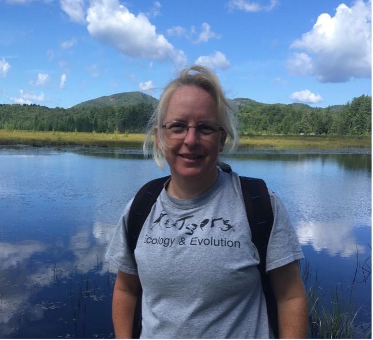 Julie Lockwood in a t-shirt that reads Rutgers Ecology & Evolution. Behind her is a lake, forested mountains, and a blue sky with white clouds.