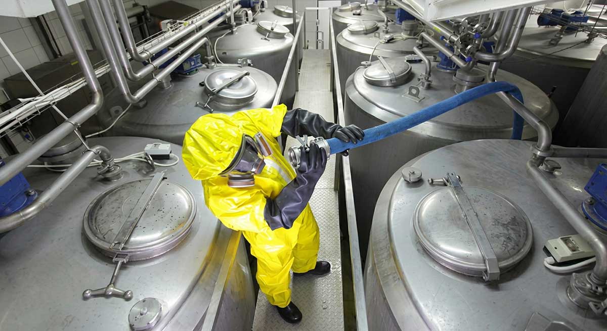 A photo of a worker in a yellow hazmat suit looking into a hose near metallic silos