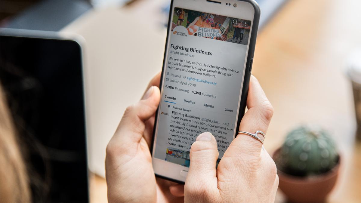 An image of a woman holding a phone. On the phone screen is the Fighting Blindness Twitter page.