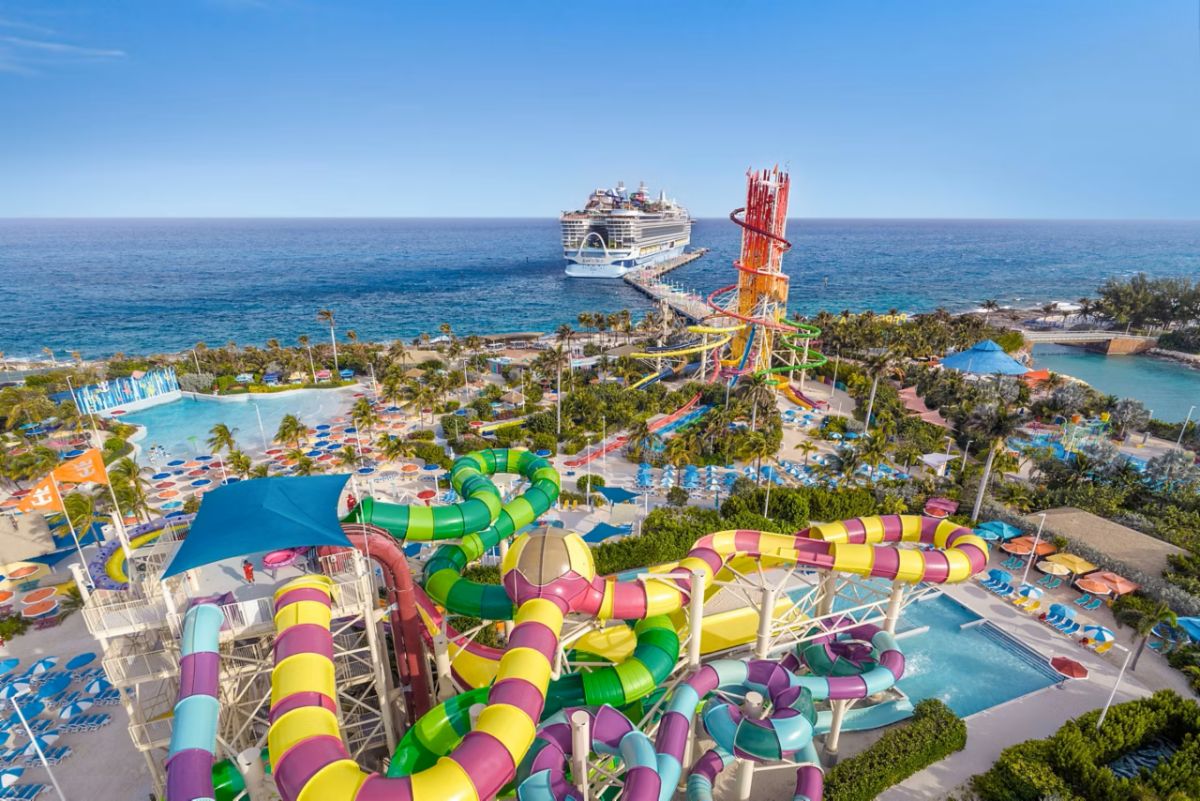 Aerial photograph showing a colorful seaside water park with intertwined green, yellow, pink, and purple slides, pools, palm trees, and lounge areas. Cruise ship offshore and tall red-orange slide tower highlight resort setting and major attractions against a blue ocean backdrop.