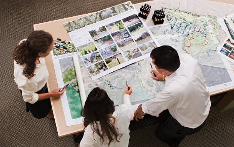 Three planners looking at blueprints and maps laid out on a table. 