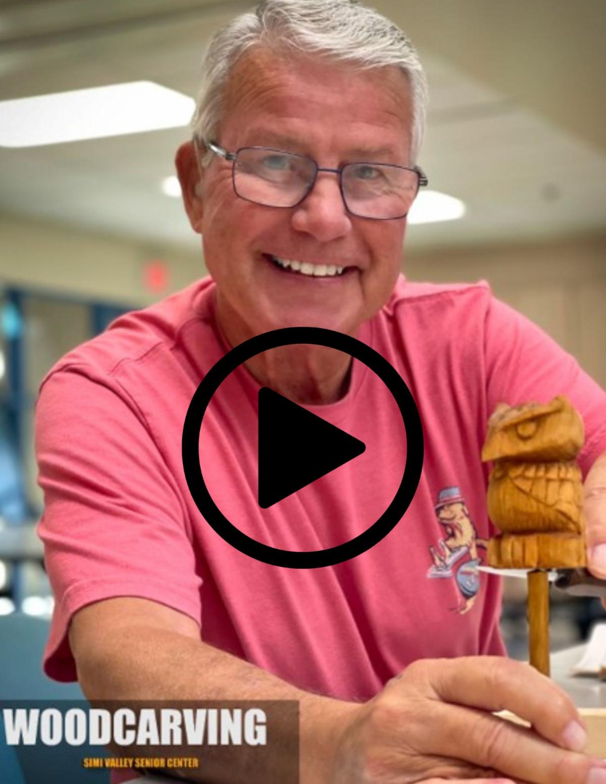 Video play button over screenshot of man in a pink shirt showing his woodcarving class project