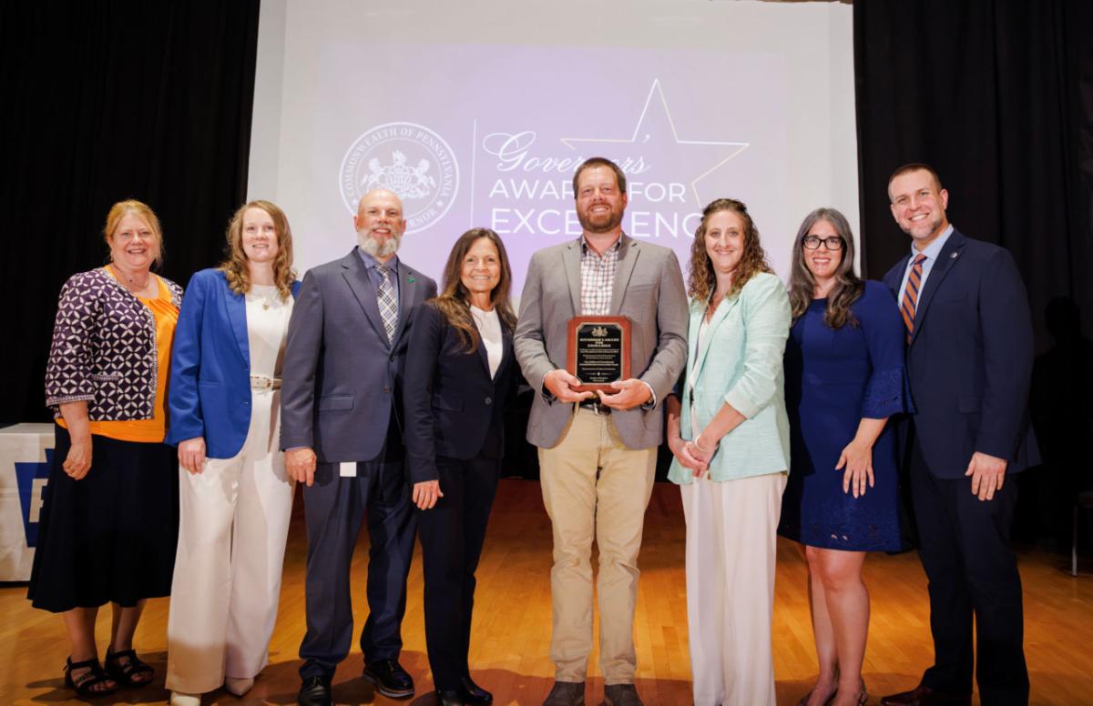 A group of seven L and I employees pose on stage with L and I Secretary Nancy A Walker with one man holding a plaque A screen in the background displays Governors Awards for Excellence