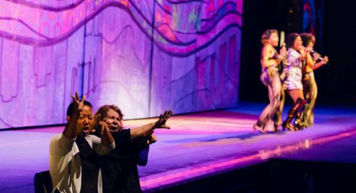 Two ASL interpreters in the foreground enthusiastically sign toward the audience while performers sing and dance onstage under colorful lighting in the background.