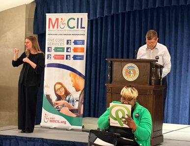 Speaker stands at a podium on a stage while a sign language interpreter gestures beside a MOCIL banner; an attendee in a green jacket sits in front, holding a folder.