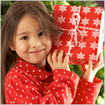 Smiling pajama-clad little girl holding a wrapped gift in front of a Christmas tree