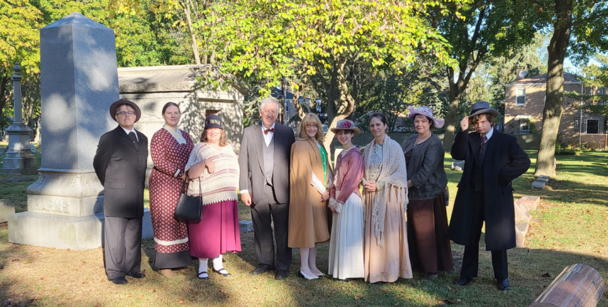 A group of volunteer actors in period costumes ranging from the mid-1800s to the mid-1900s stand in a cemetery smiling for the camera.