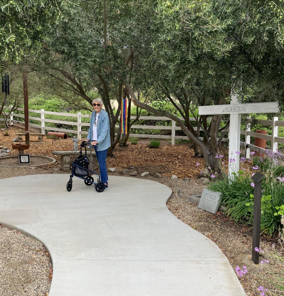 A woman stands with her walker on the concrete path with olive trees and a white fence behind her and a cross and bench closer.