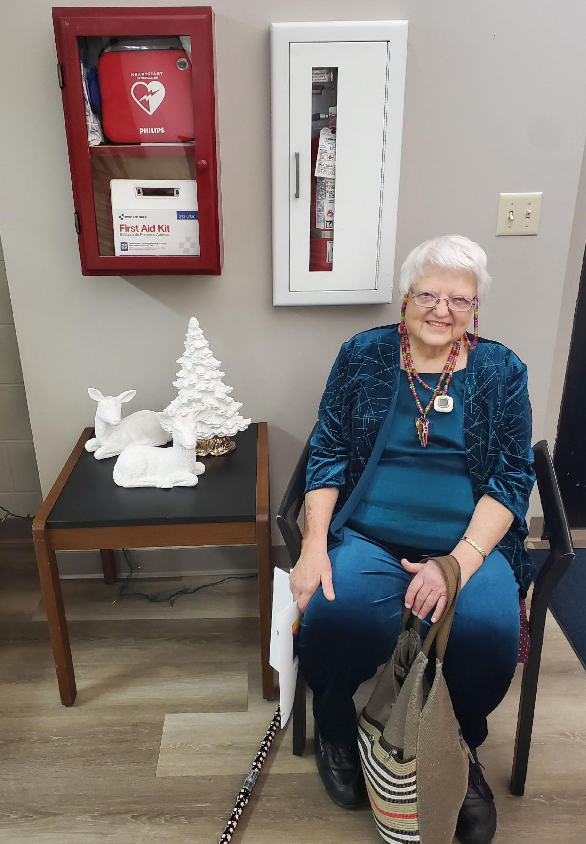A smiling white-haired woman in blue sits in a chair with armrests next to ceramic Christmas decorations. A fire extinguisher and AED are on the wall behind her..m