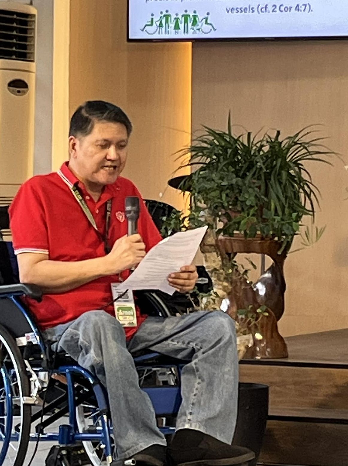 A man with black hair and red shirt who is seated in a wheelchair holds a microphone reads from a paper