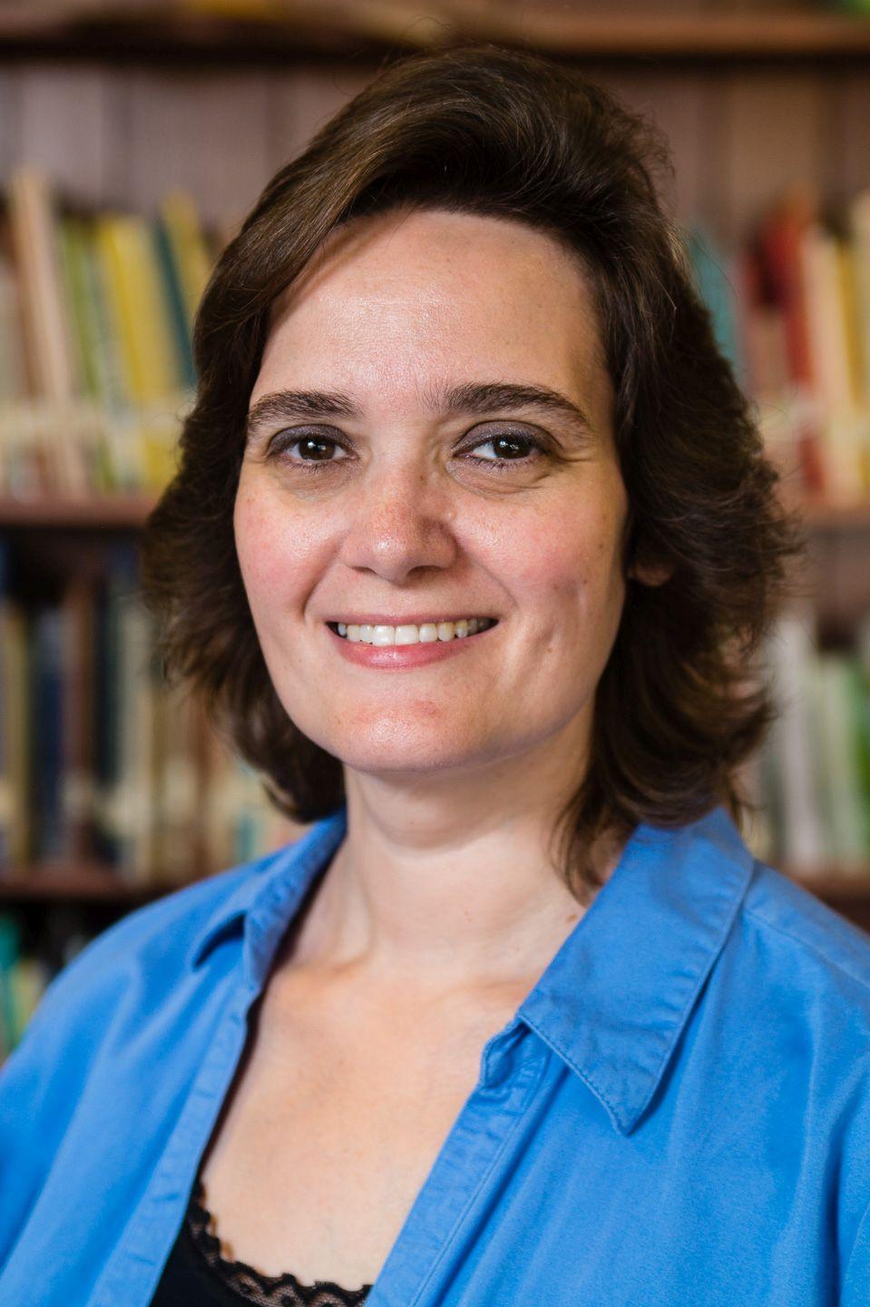 Smiling Caucasian woman with brown collar length hair and blue shirt_ bookshelf is behind her