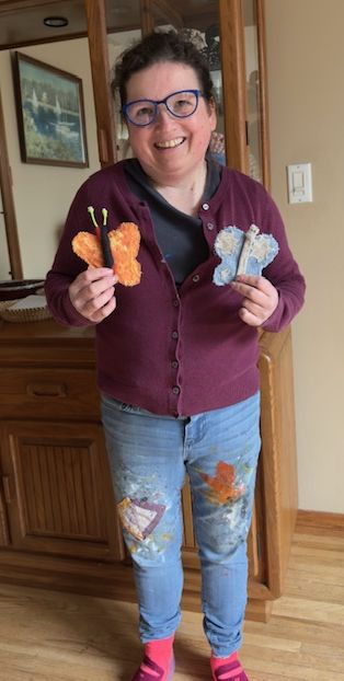 Short smiling Caucasian woman wearing glasses holds two fabric butterflies.