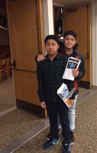 Two youth stand at sanctuary entrance holding bulletins. The elder brother has his arm around his brother. 