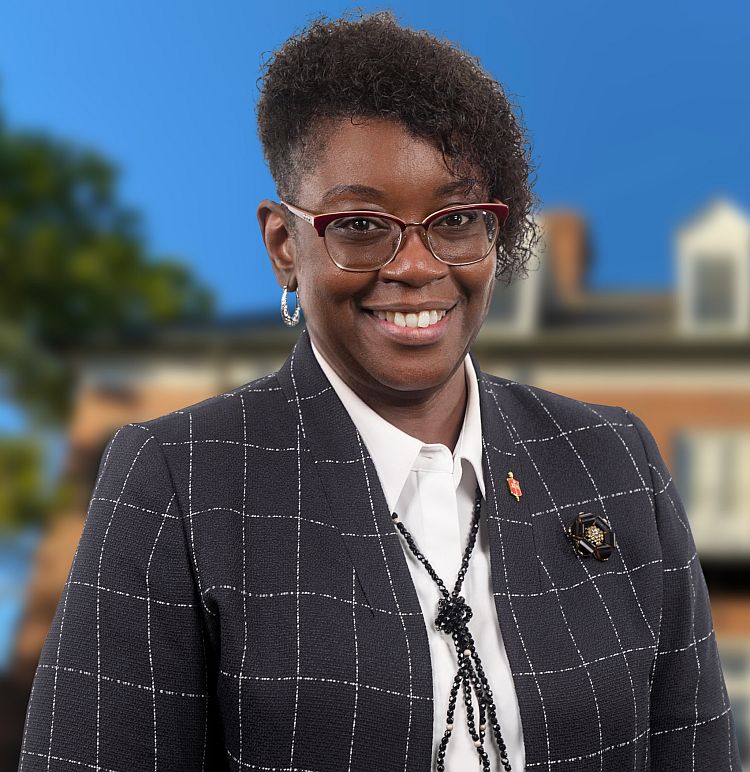 A smiling black woman with short hair and glasses with a campus setting in the background