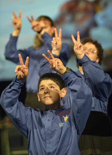 Three choir member in blue shirts hold both hands up with fingers making a V shape