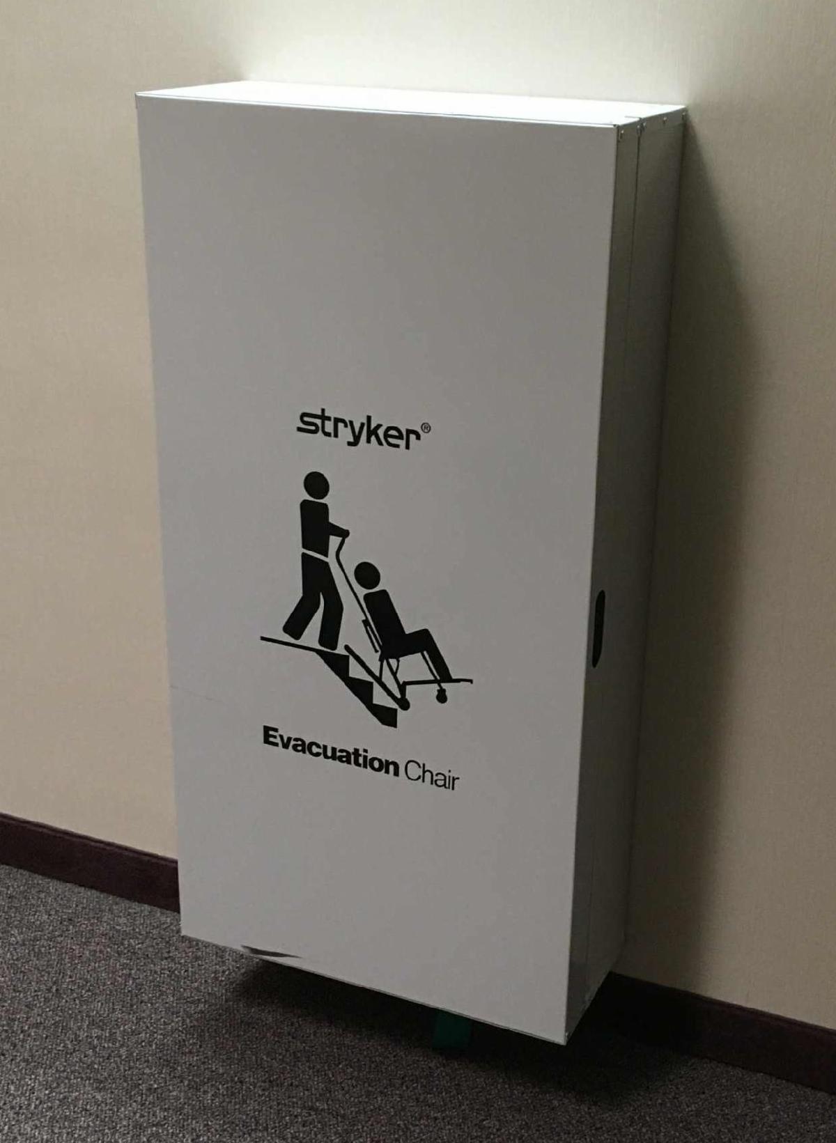 A large grey wall-mounted box has an image of a person assisting another using an evacuation chair. The brand Stryker is above the image.