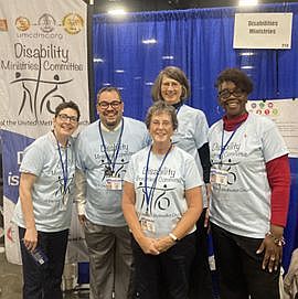 Four women and one man wearing light blue Disability Ministries Committee T-shirts stand in front of the committee banner
