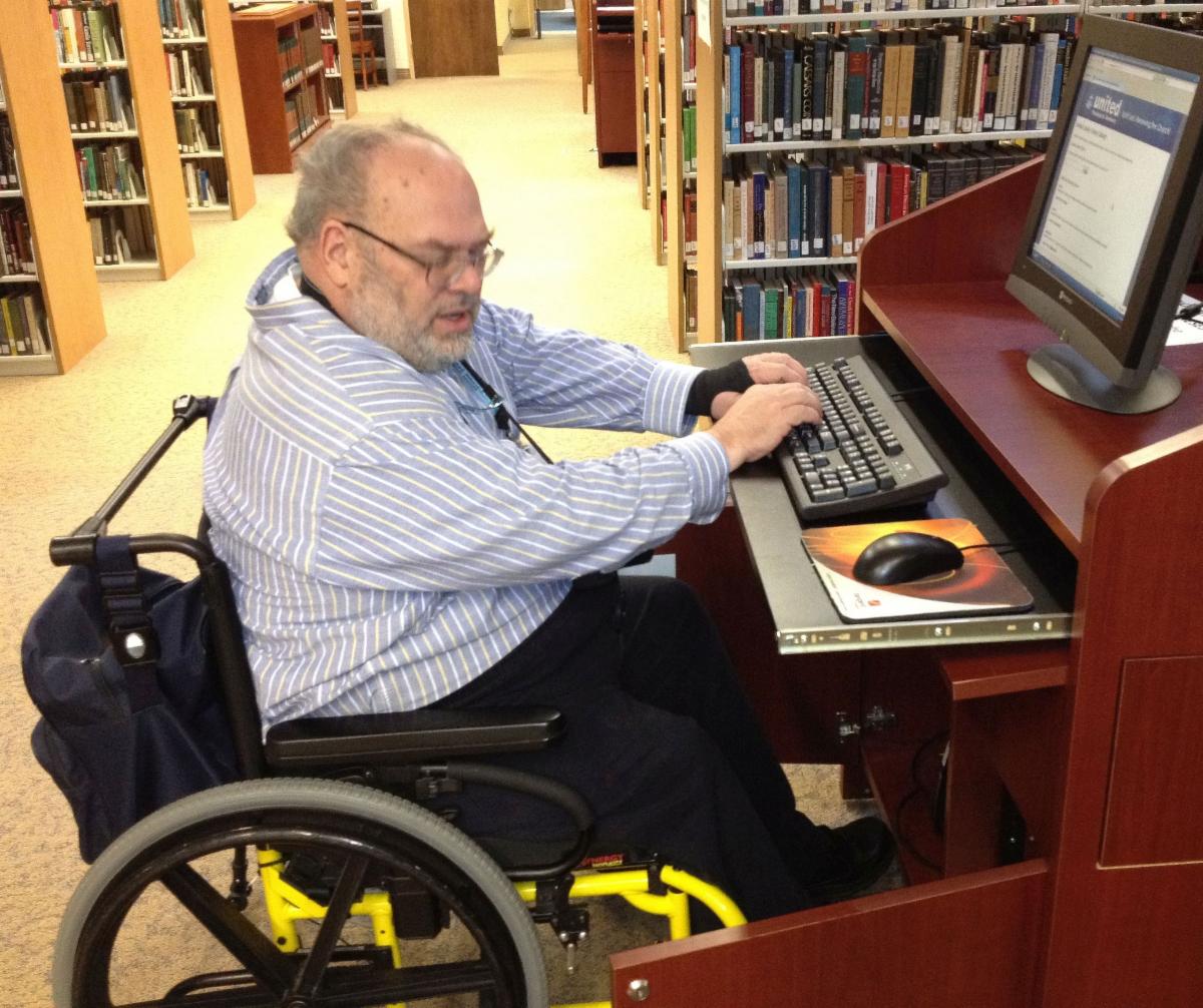 A man using a wheelchair and wearing a blue striped shirt and glasses sits at a computer with library bookshelves in the background.