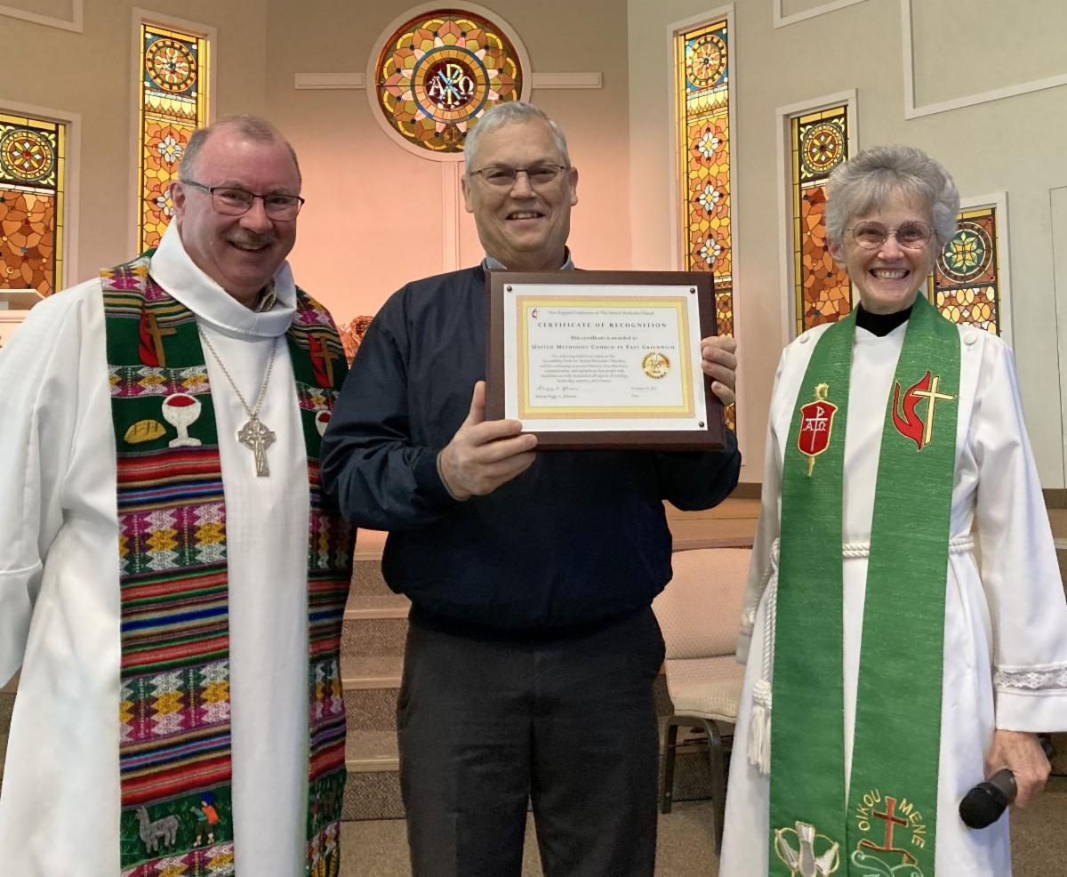 Two pastors in white robes flank the trustee holding the gold badge certificate. Six stained glass windows are in the background.