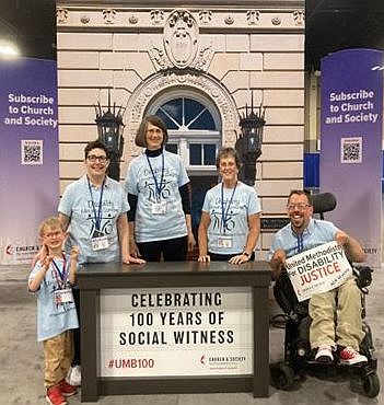 A child, three standing women, and a man seated in a wheelchair are in front of the replica UM building behind a sign celebrating 100 years of social witness