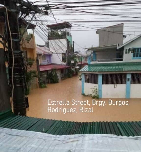 A street between buildings looks like a muddy brown river.  Many electrical wires are stretched over the buildings.