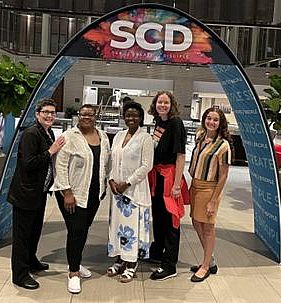 Five smiling women of various skin tones stand under a display arch with SCD over their heads