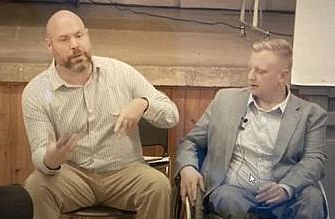 Two seated middle aged men: one speaks and gestures to the audience, the other who uses a wheelchair looks at him. A stage is in the background.                               and one in a wheelchair 