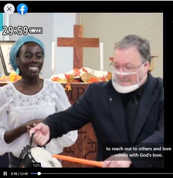 Screenshot shows worship leader Naomi in blue scarf and while blouse smiling while Pastor Tom in clerical garb and face mask with transparent mouth piece hitting a drum