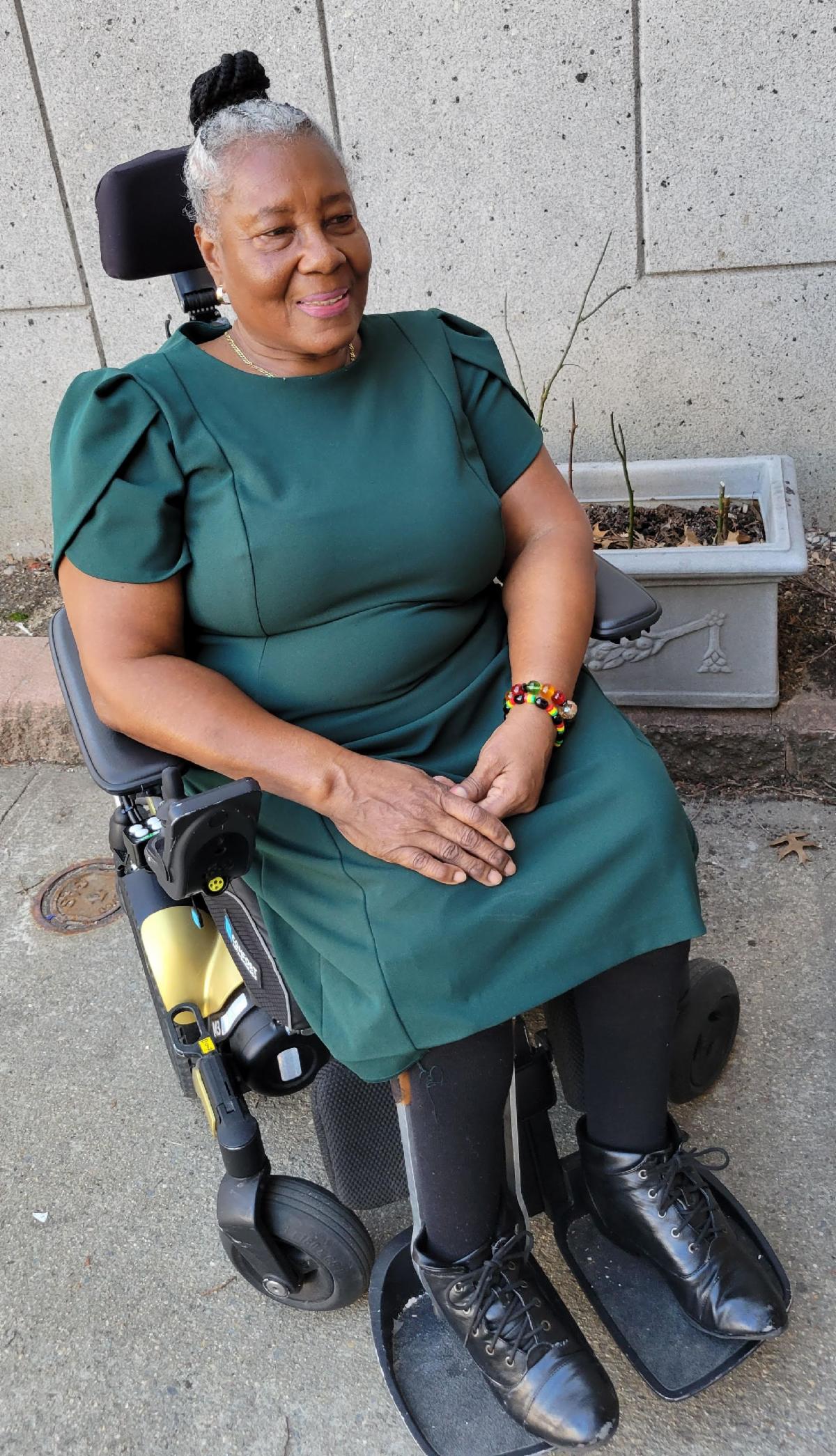 A smiling older black woman in green dress and leg braces showing sits in her wheelchair.