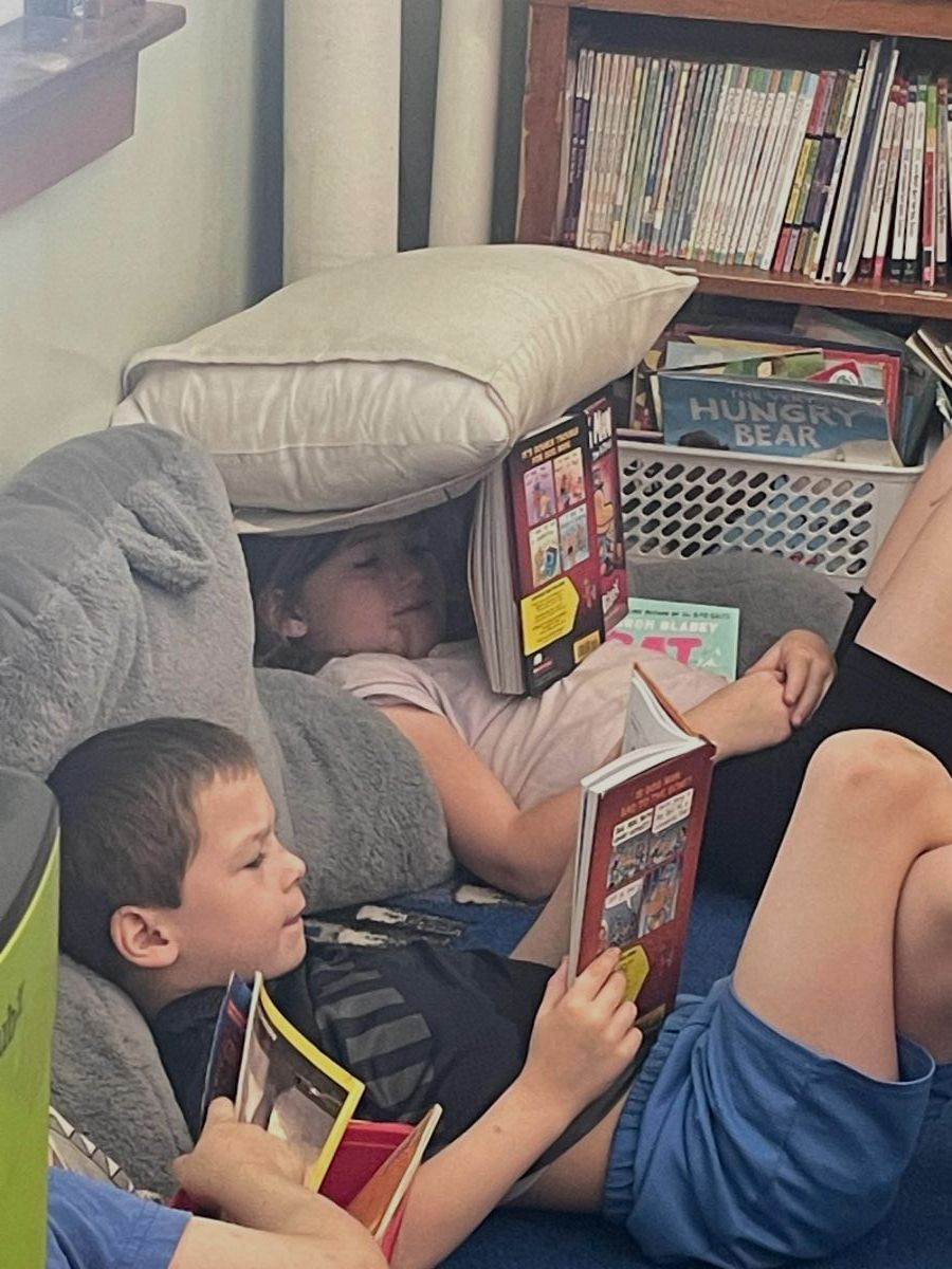 Two young students are sitting on the floor, reading a book. One has a pillow over their head.