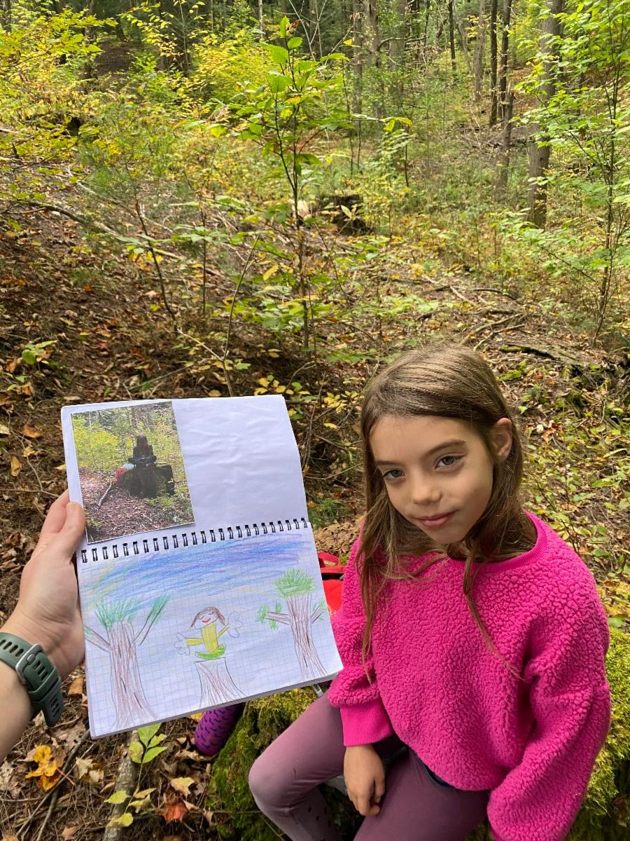 A young student is sitting outside in a forest. Off-camera, an adult is holding up a notebook showing a photograph, and the student's drawing of what's in the photograph.