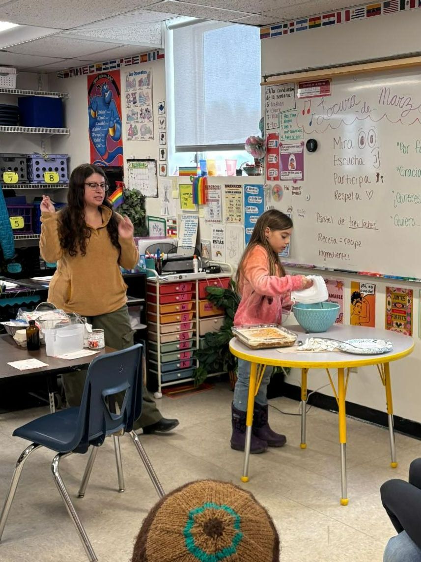 A teacher sits on the floor with students, looking at papers lined up on the ground.
