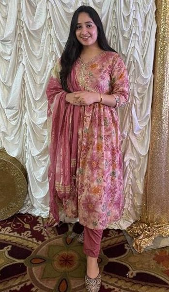 A student stands in front of an ornate draped backdrop wearing a pink and gold traditional outfit, smiling at the camera.