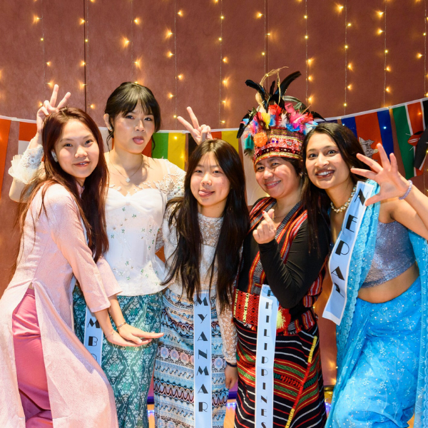 Five international students pose together in colorful traditional clothing in front of string lights and flags, smiling and making playful peace signs.