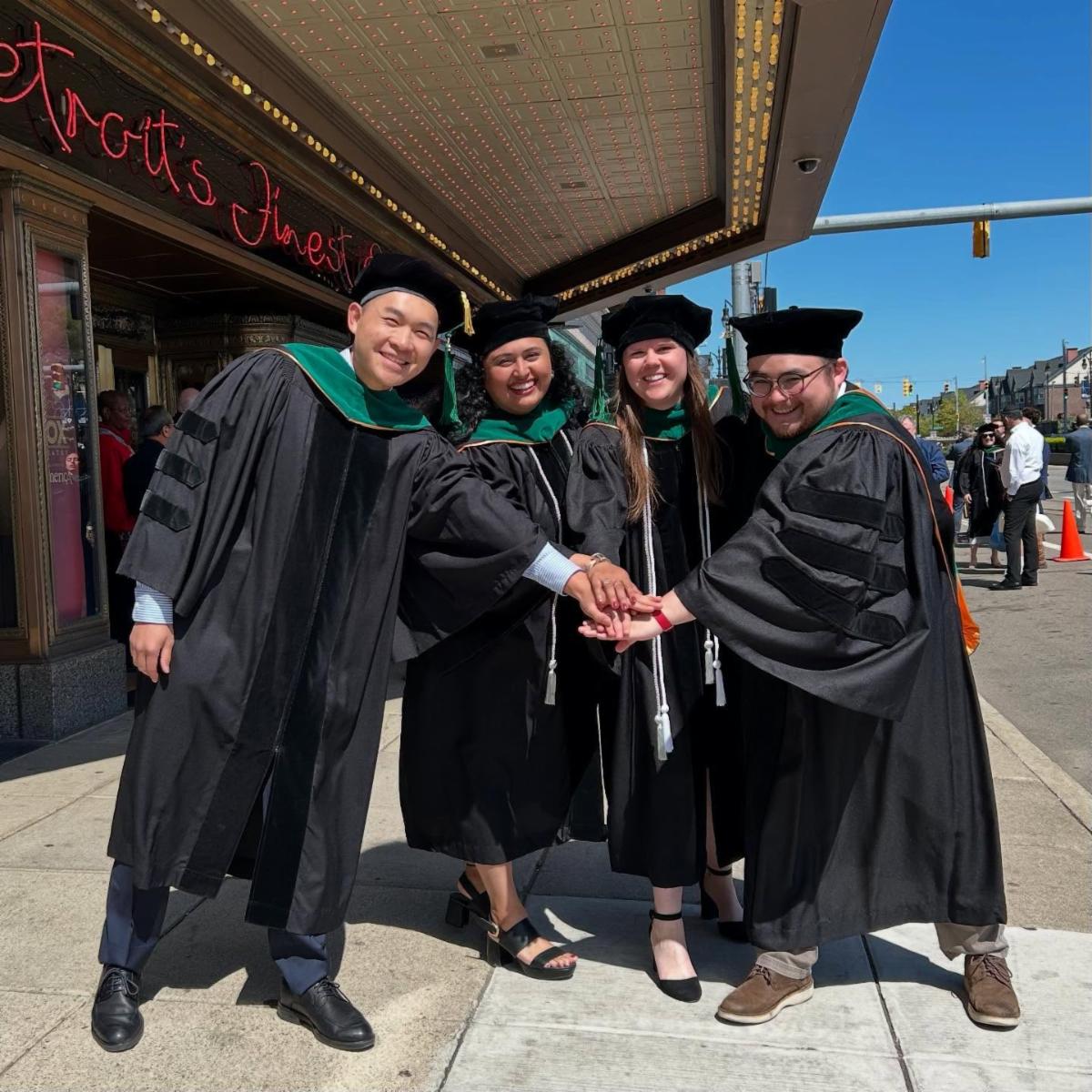 Four medical students stand with their hands in the center in graduation regalia just before Wayne State University School of Medicine Class of 2025 graduation at the Fox Theater in Detroit.