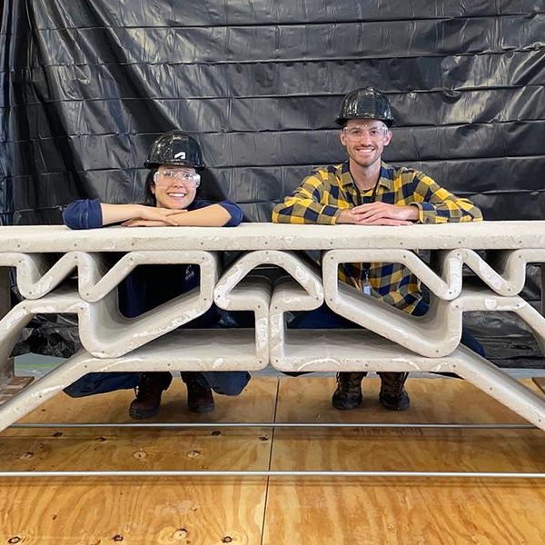 Hajin Kim-Tackowiak and Zane Schemmer, in hard hats and safety glasses, kneel behind a concrete bridge about the size of a park bench.