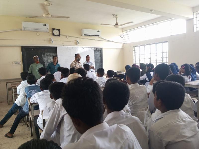 Small classroom with roughly 35 young students in uniforms sitting facing the blackboard and four teachers. Two mini-split AC units are visible above the chalkboard.