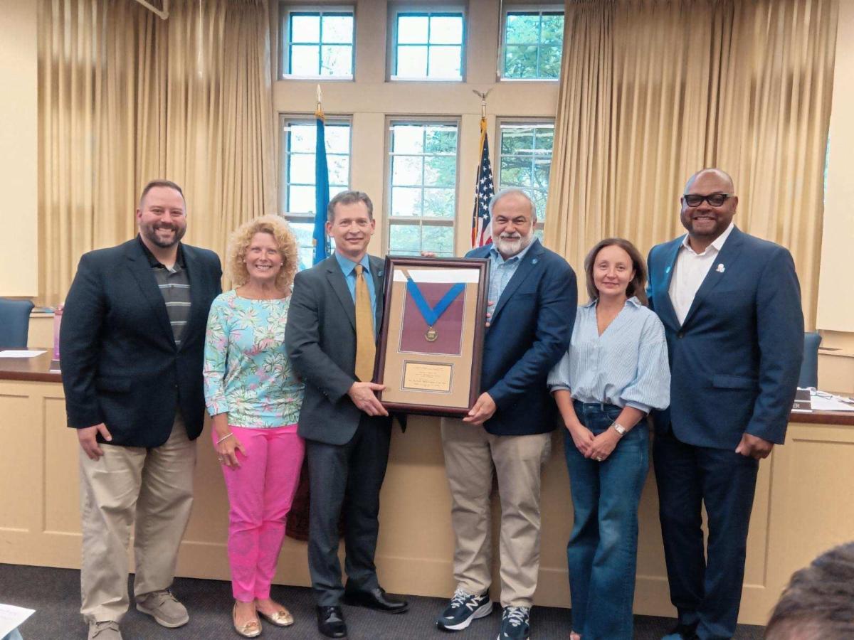 A group of six professionally dressed individuals pose indoors in front of a large window and American flags. The person in the center holds a framed award or plaque with a blue ribbon and gold medallion. The setting appears to be a formal recognition or a