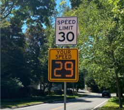 Speed feedback sign displaying a driver’s speed of 29 mph below a 30 mph speed limit sign on a tree-lined residential street