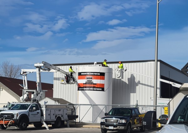 workers in safety gear using bucket trucks to apply signage on a large outdoor tank at a public works facility with clear blue sky above