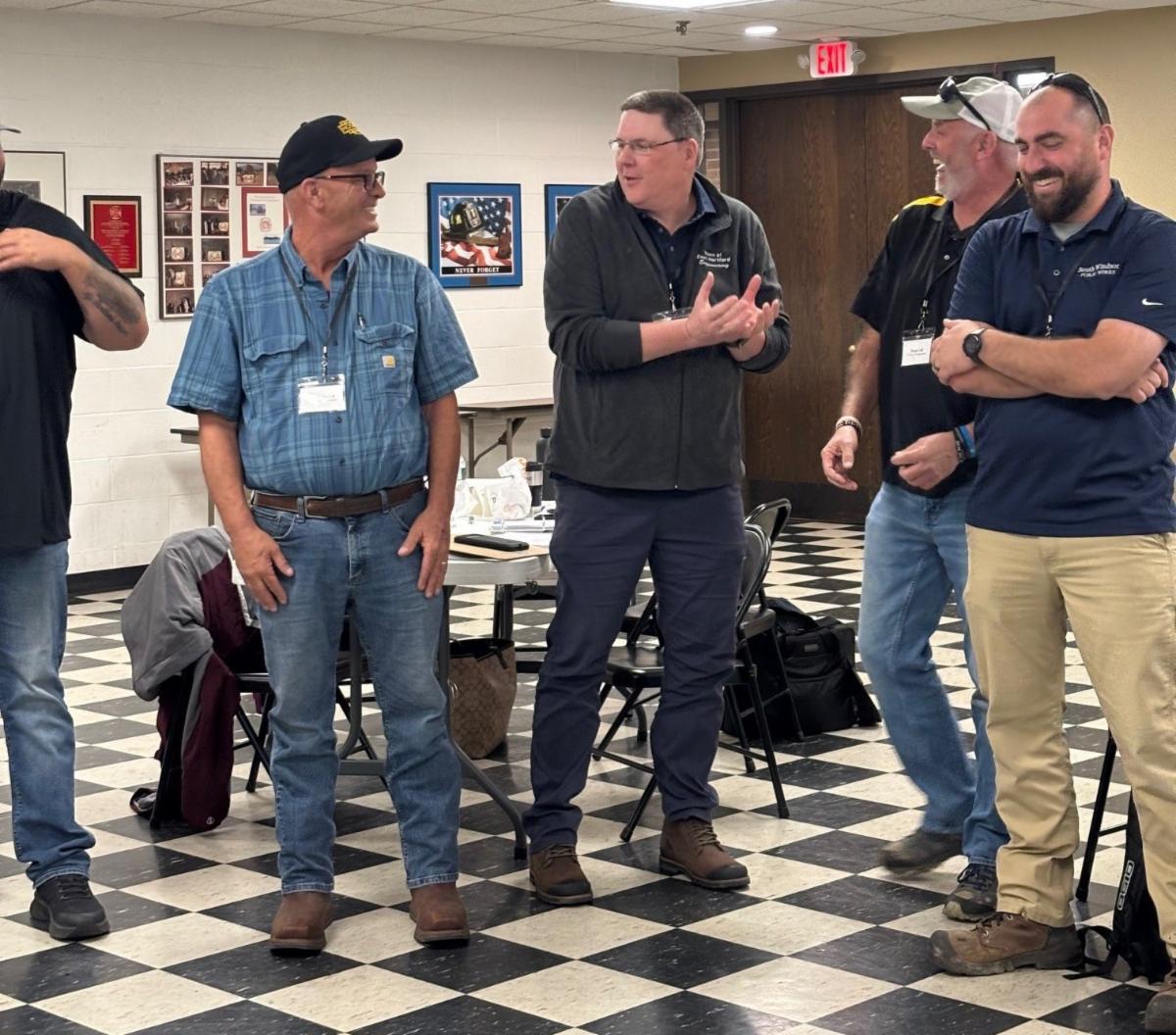 five men standing and talking in a classroom with black and white checkered floor wearing name badges and casual clothes smiling and laughing together