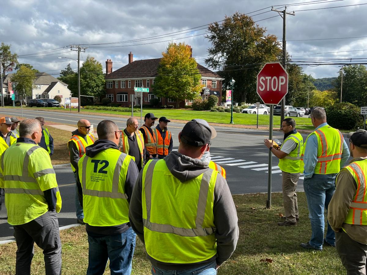 A group of public works professionals wearing high-visibility safety vests gathers around an instructor holding a stop sign during an outdoor roadway safety training. The session takes place at an intersection near a residential neighborhood under a partly cloudy sky.