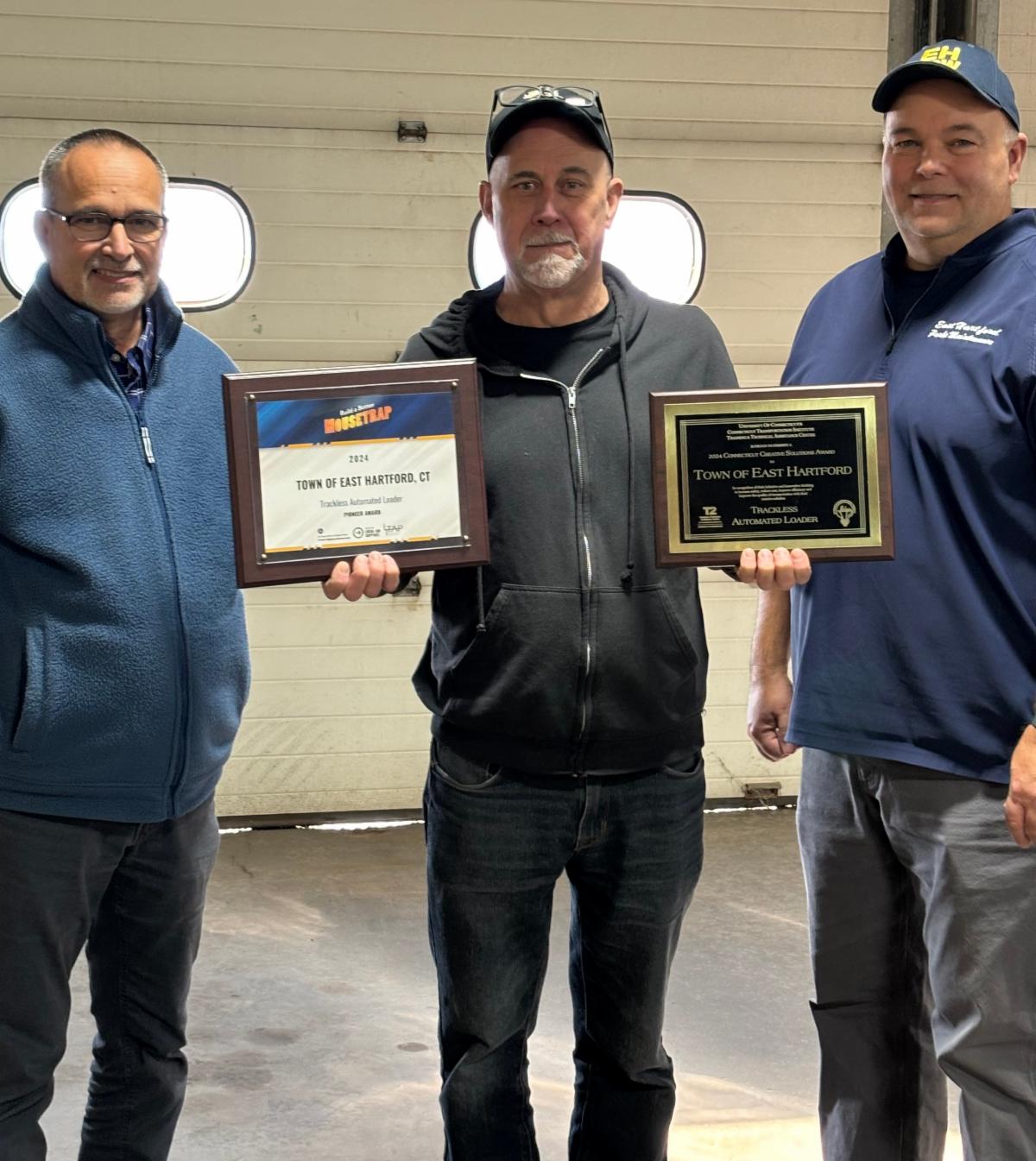 Three men standing indoors holding award plaques, recognizing the Town of East Hartford for a trackless automated loader project.