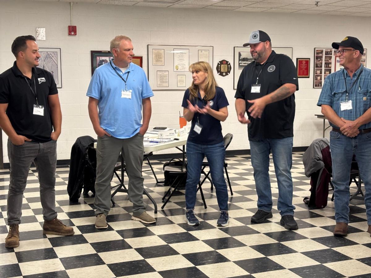 five people standing in a classroom with black and white checkered floor two are speaking while others listen with tables and framed items on the wall in the background