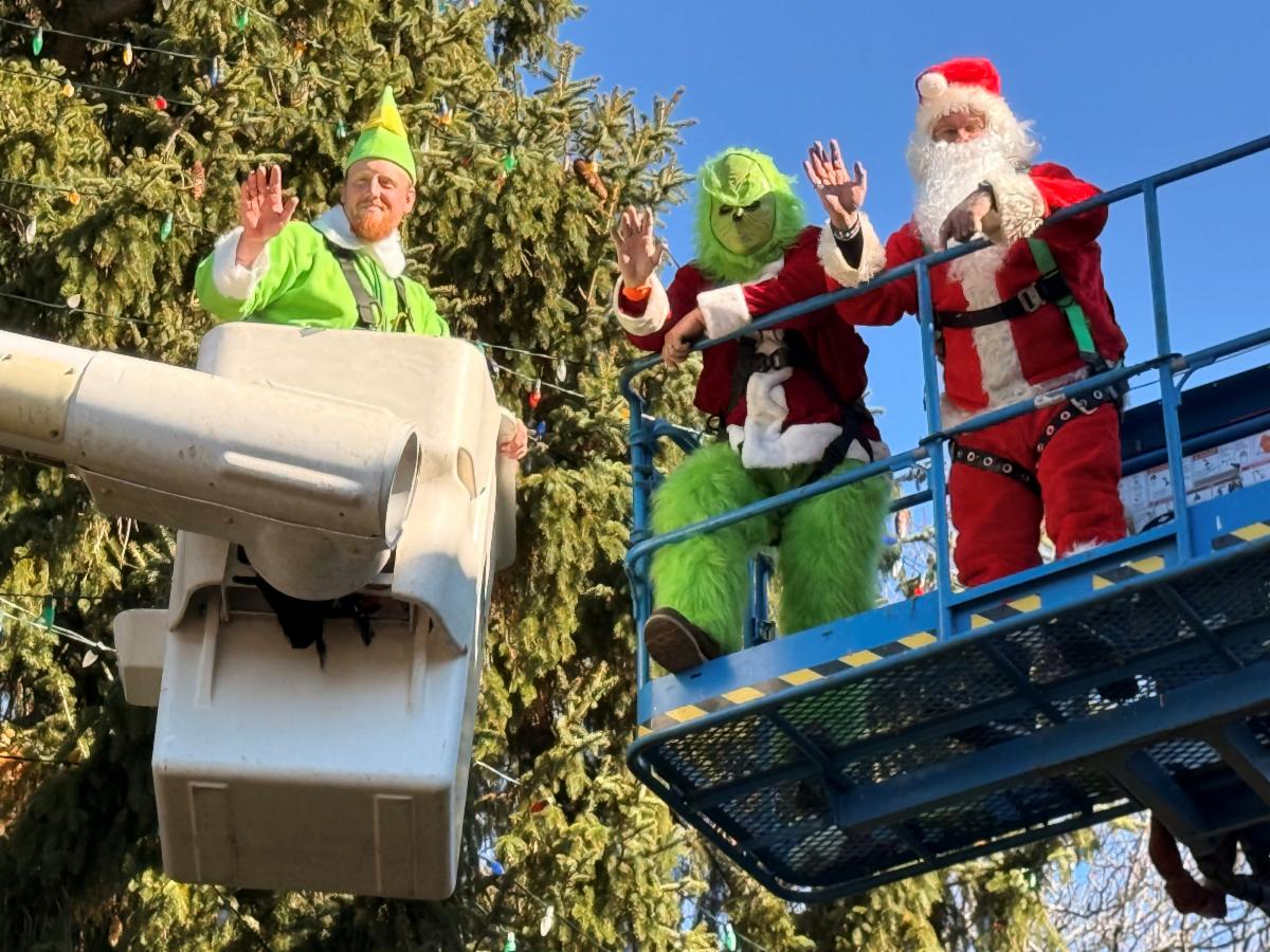 three people dressed as Buddy the Elf the Grinch and Santa Claus waving from two elevated work platforms in front of a large outdoor Christmas tree on a sunny day
