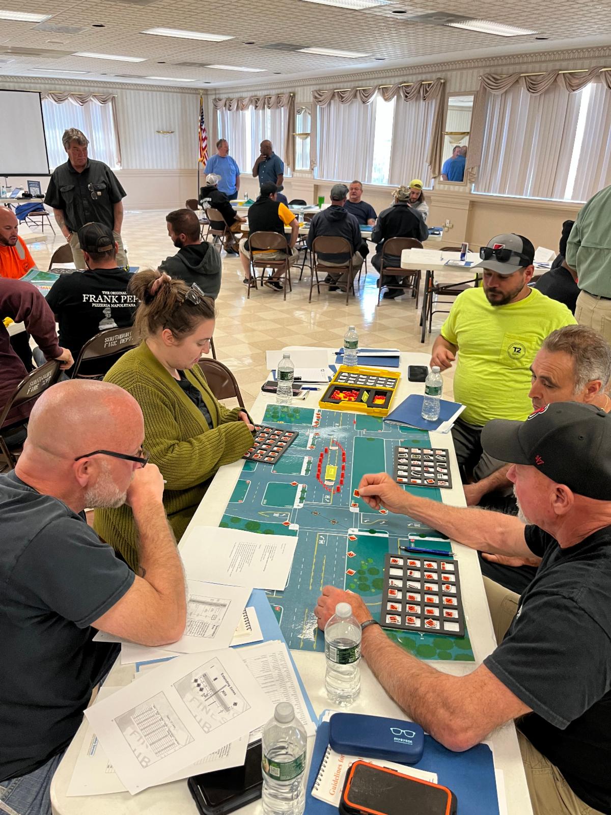 Participants sit around a table in a large room, actively engaged in a hands-on training exercise using a tabletop traffic layout and various instructional tools. Other groups work at nearby tables, contributing to a collaborative workshop environment.