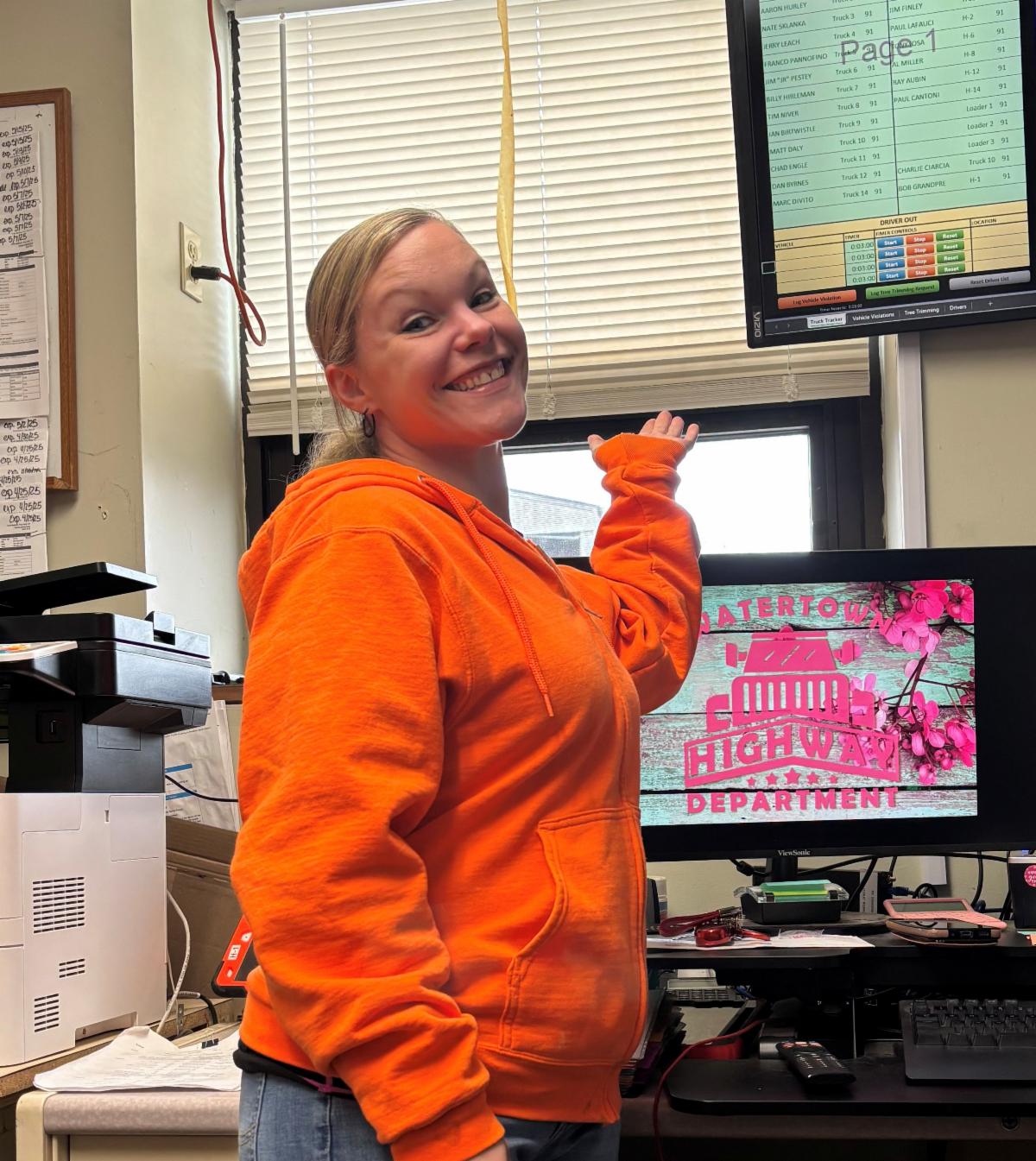 Woman in an orange hoodie smiles and gestures toward a screen displaying a list of names and vehicles. Another monitor shows a pink and red Watertown Highway Department logo with flowers.