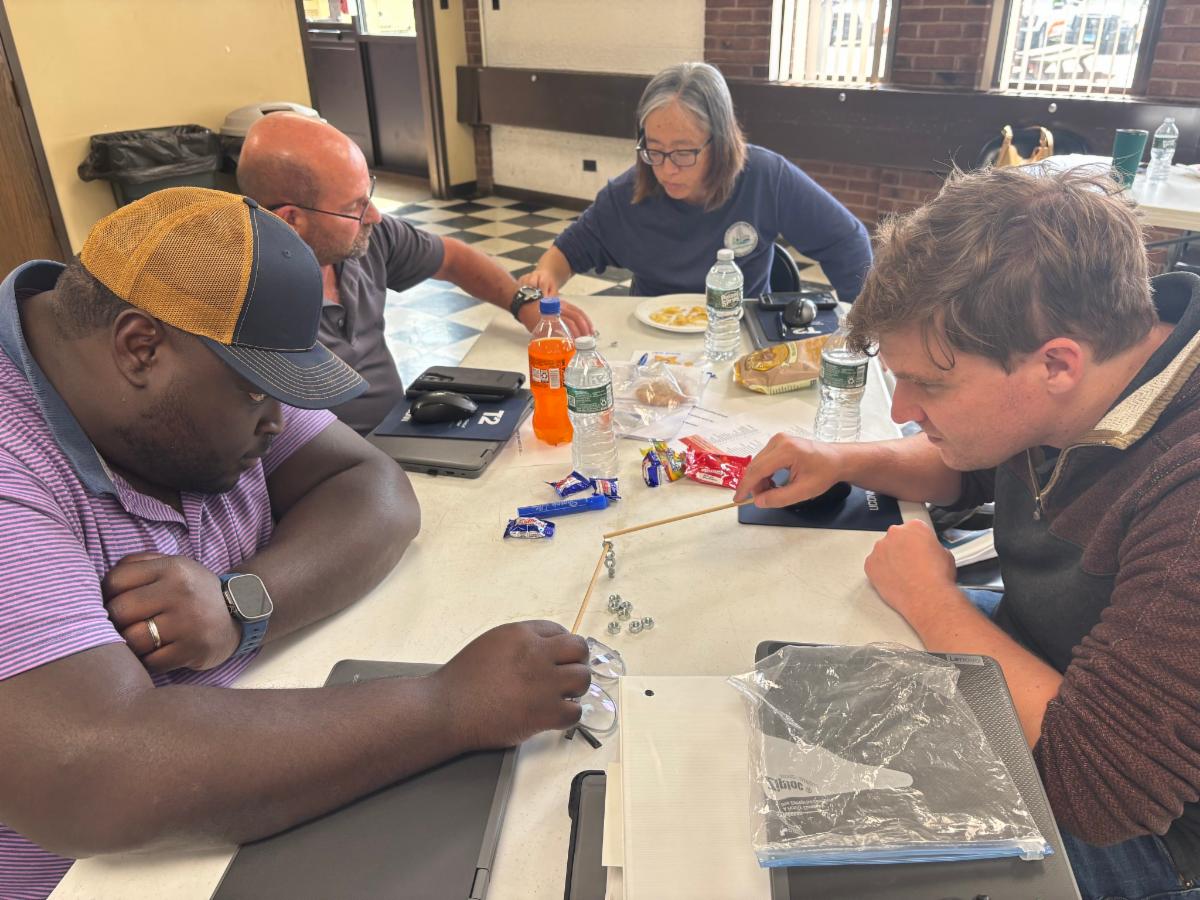 four people sit around a table working on a team activity two men focus on stacking metal nuts using wooden sticks while the others watch snacks water bottles laptops and notebooks are on the table the group appears engaged and concentrated in a classroom 
