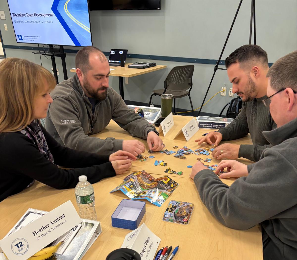four people sitting around a table working together on a puzzle during a workplace team development session with a presentation screen in the background